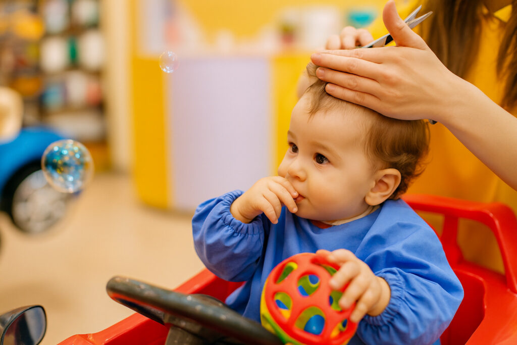 Малыш сидит в детском кресле-машинке во время стрижки