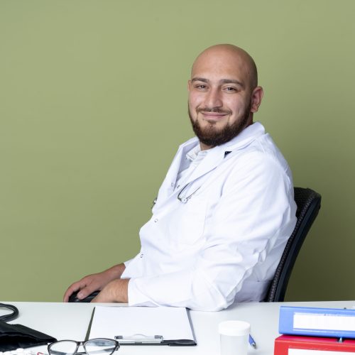 pleased young bald male doctor wearing medical robe and stethoscope sitting at desk work with medical tools isolated on green background